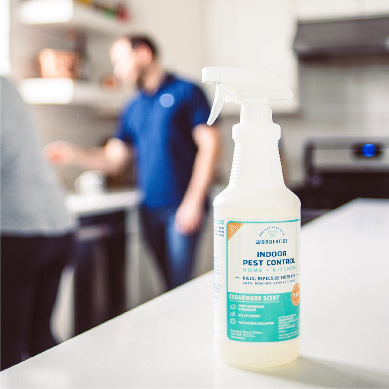 A 128 oz. Wondercide Cedarwood Indoor Pest Control spray sits on a kitchen counter, its label showing it's safe for pets—perfect for homes with furry friends—as two people are blurred in the background near the stove.