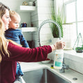 A woman uses Wondercide 128 oz. Cedarwood Indoor Pest Control, safe around pets, as she sprays her kitchen sink by a sunny window with plants, holding her toddler and enjoying the fresh cedarwood scent.