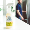 A 128 oz. bottle of Wondercide Lemongrass Indoor Pest Control rests on a white countertop, as a blurred person in a blue shirt opens the refrigerator in the background.