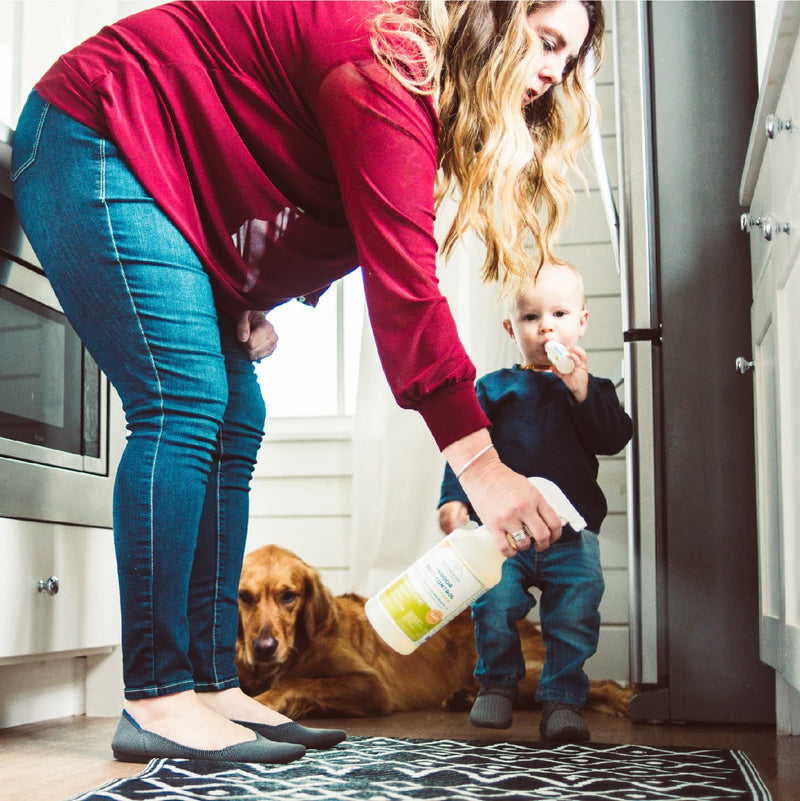 A woman sprays Wondercide’s 128 oz. Lemongrass Indoor Pest Control on her kitchen floor, while a toddler enjoys a snack and a golden retriever relaxes nearby in the bright, pet-safe space with white cabinets.