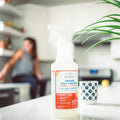 A 128 oz. bottle of Wondercide Peppermint Indoor Pest Control sits on a white kitchen counter beside green plant leaves, a patterned mug, and a blurred person—safe for pets and effective for indoor pest protection.