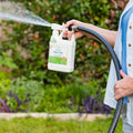 A person uses Wondercide Flea & Tick Yard Spray Refill Starter Kit, spraying water in a garden with lush green grass and plants to control fleas and ticks effectively.