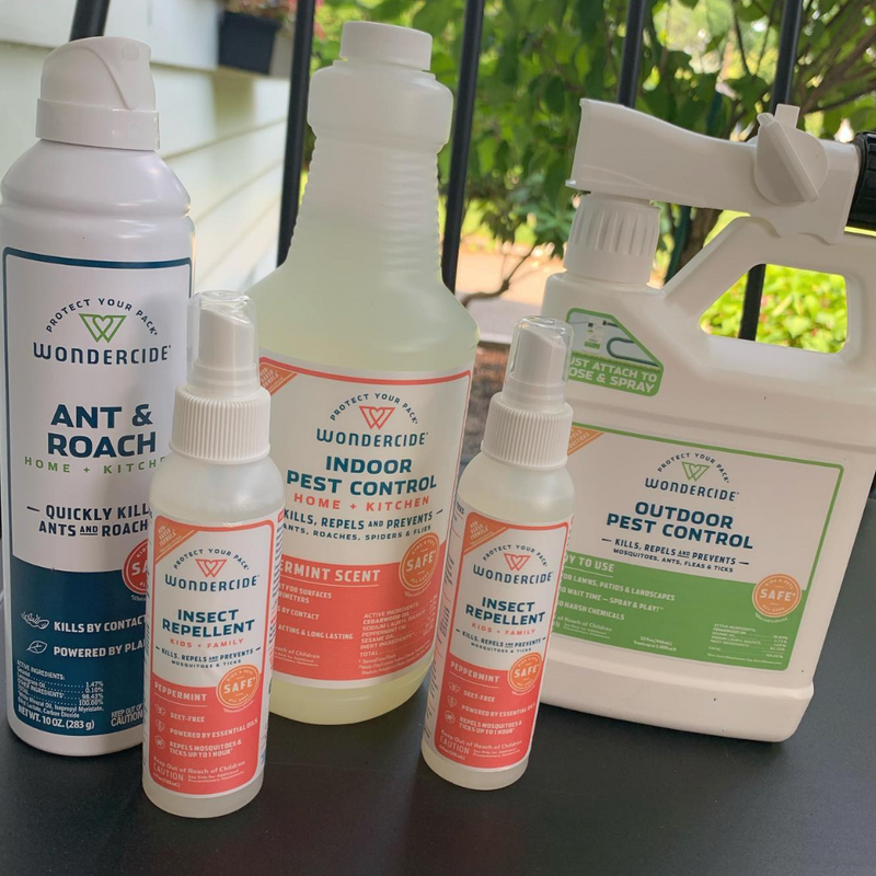 Bottles of various Wondercide pest control products are arranged on a table. Visible are Ant and Roach spray, Indoor Pest Control, Insect Repellent, and Outdoor Pest Control. The setting appears to be an outdoor area with a porch and greenery.