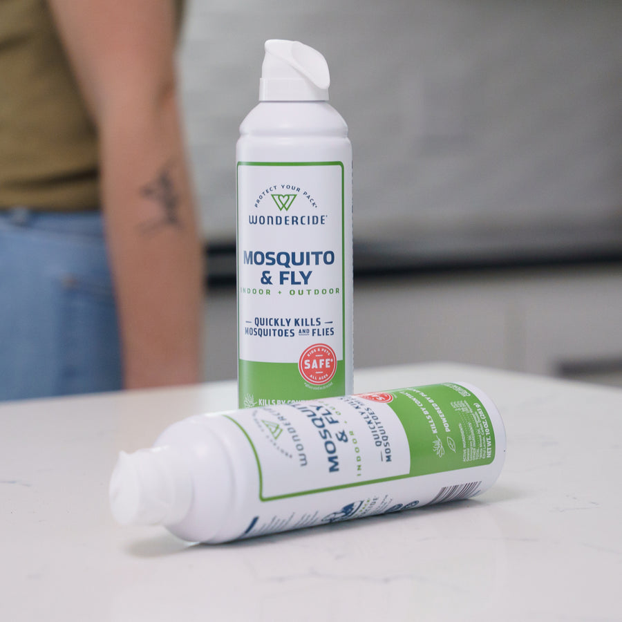 Two cans of Wondercide Mosquito & Fly for Indoor + Outdoor spray are on a white countertop—one upright, one lying down. A tattooed arm is blurred in the background. This plant-powered spray is pet-safe and effectively kills mosquitoes.