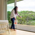 A woman in a white shirt and jeans uses Wondercide's Home Treatment Kit | Flea & Tick Spray from a large container on the floor near a glass door, with a scenic green home and yard visible in the background.