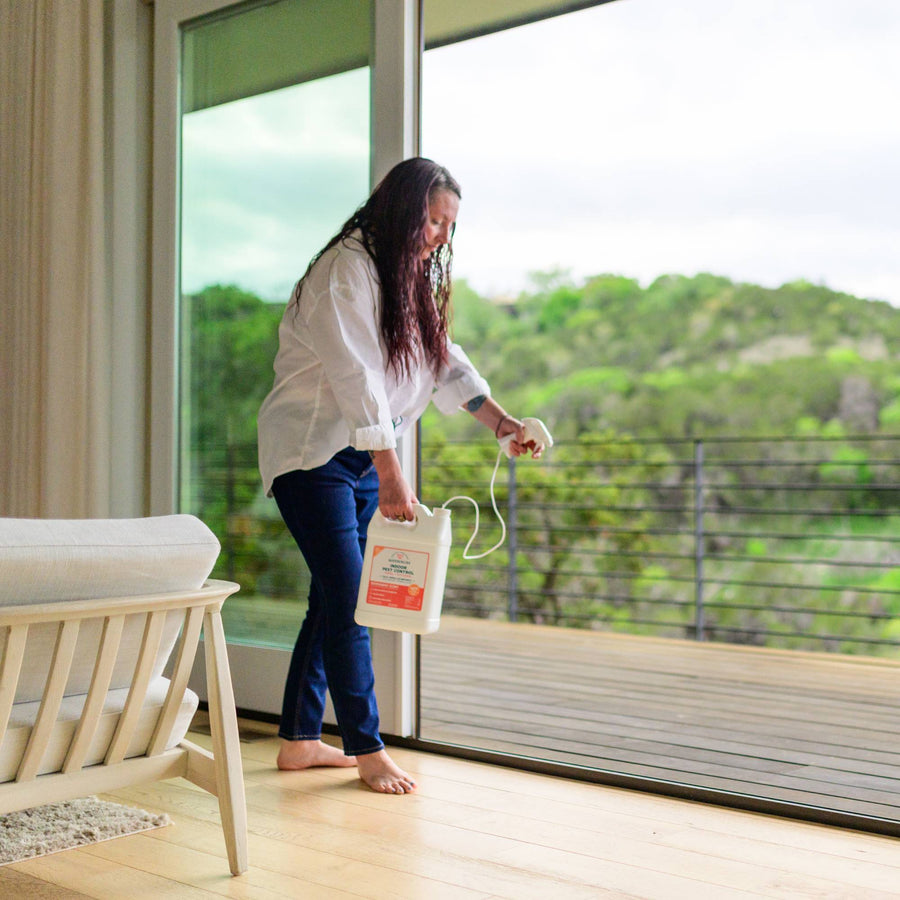 A woman in a white shirt and jeans uses Wondercide's Home Treatment Kit | Flea & Tick Spray from a large container on the floor near a glass door, with a scenic green home and yard visible in the background.
