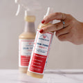 A hand with red nail polish holds Wondercide Skin Tonic Itch Spray for pets, formulated to soothe itching, with another Wondercide spray bottle blurred in the background on a light surface.