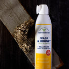 A can of Wondercide Wasp & Hornet for Porch + Patio stands upright beside a wasp nest on wood, both on a dark surface. The can features plant-based, family-safe, and effective wasp and yellow jacket killer benefits.