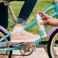 A hand applies Wondercide Cedarwood Insect Repellent for Family to a child's ankle, wearing pink sneakers and white socks on a blue and purple bike, with grass in the background.