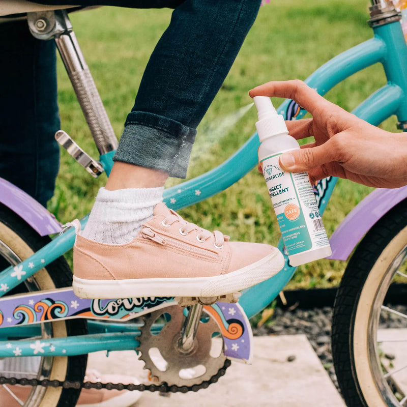 A hand applies Wondercide Cedarwood Insect Repellent for Family to a child's ankle, wearing pink sneakers and white socks on a blue and purple bike, with grass in the background.