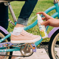 An adult sprays Wondercide Lemongrass Insect Repellent for Family on a child's ankle as the child, in pink sneakers, white socks, and rolled-up jeans, sits on a blue bike with purple and pink accents. Grass can be seen in the background.