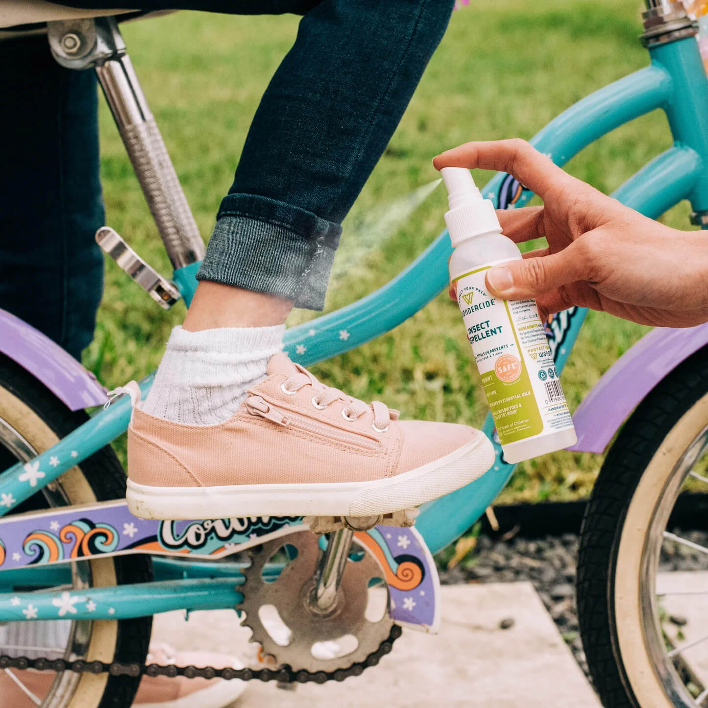 A person applies Wondercide Lemongrass Insect Repellent for Family with Natural Essential Oils to a childs ankle as the child, wearing pink sneakers and rolled-up jeans, sits on a blue and purple bike surrounded by grass.