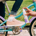 A person applies Wondercide Insect Repellent for Family to a child's ankle as the child, in pink sneakers and rolled-up jeans, sits on a blue and purple bike outdoors, helping repel ticks and mosquitoes for safe playtime near the grass.
