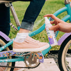 A person applies Wondercide Insect Repellent for Family to a child's ankle as the child, in pink sneakers and rolled-up jeans, sits on a blue and purple bike outdoors, helping repel ticks and mosquitoes for safe playtime near the grass.