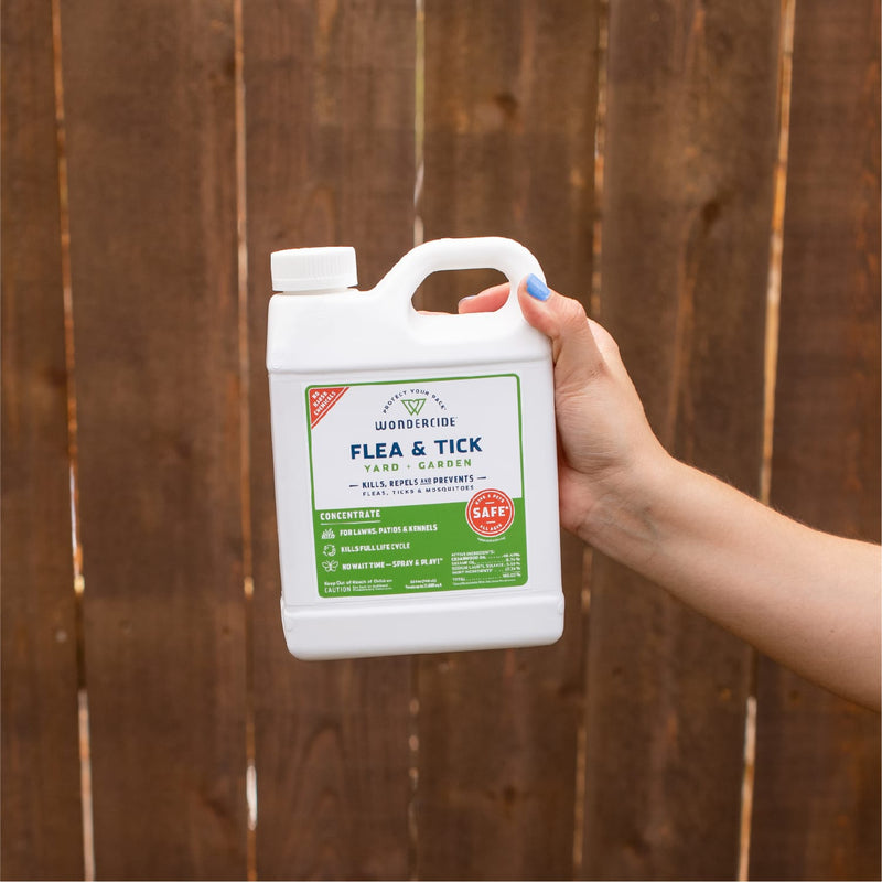 A hand with blue nail polish holds a white bottle of Wondercide Flea & Tick Concentrate for Yard + Garden (16 oz) in front of a wooden fence, highlighting easy pest protection and mosquito repellent for your outdoor space.